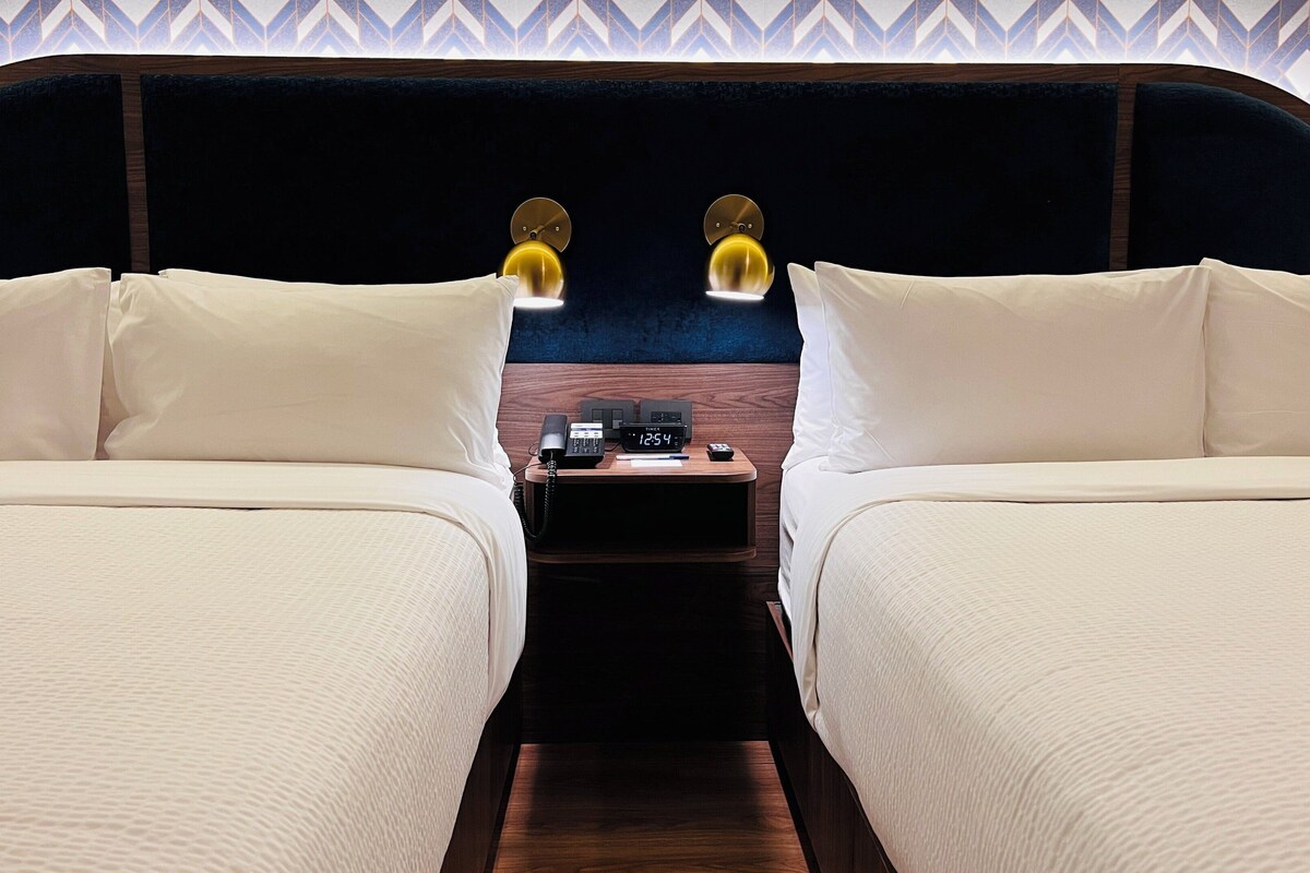 Habitación de hotel con dos camas, cabecera de madera, mesita de noche con un reloj y un papel tapiz estampado.