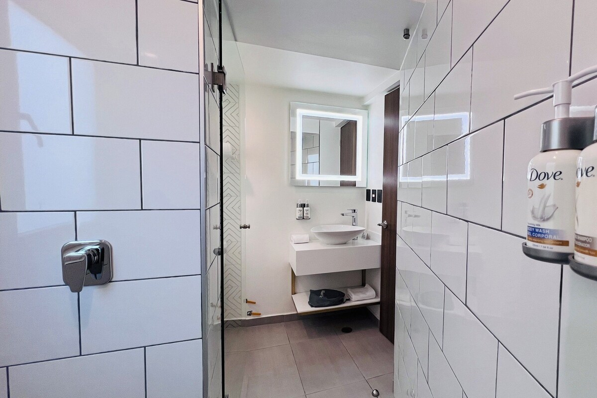 Un baño moderno con azulejos blancos, un lavamanos, un espejo y dispensadores de champú Dove.