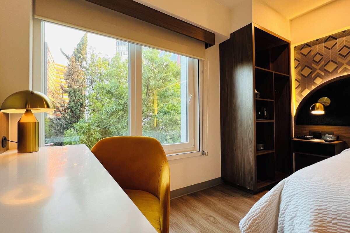 Habitación de hotel con cama, escritorio con lámpara, silla, armario y ventana con vistas a árboles.