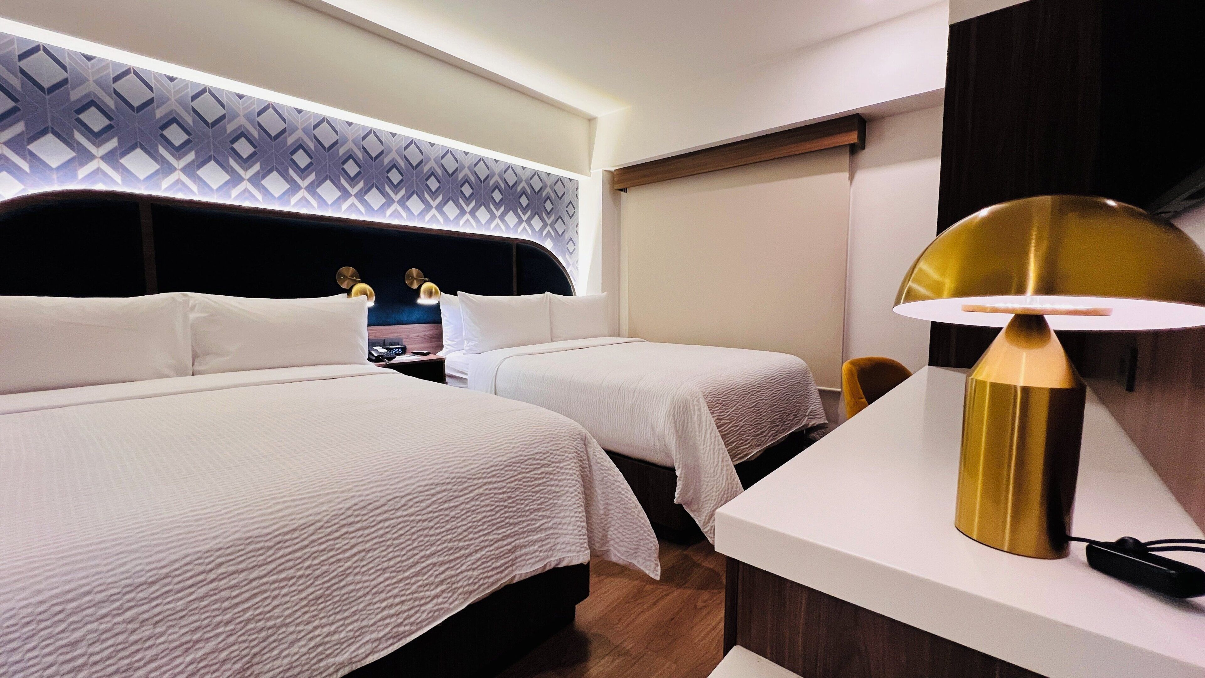 Ein Hotelzimmer mit zwei Betten, einem Schreibtisch, einer Lampe und einem gemusterten Kopfteil.