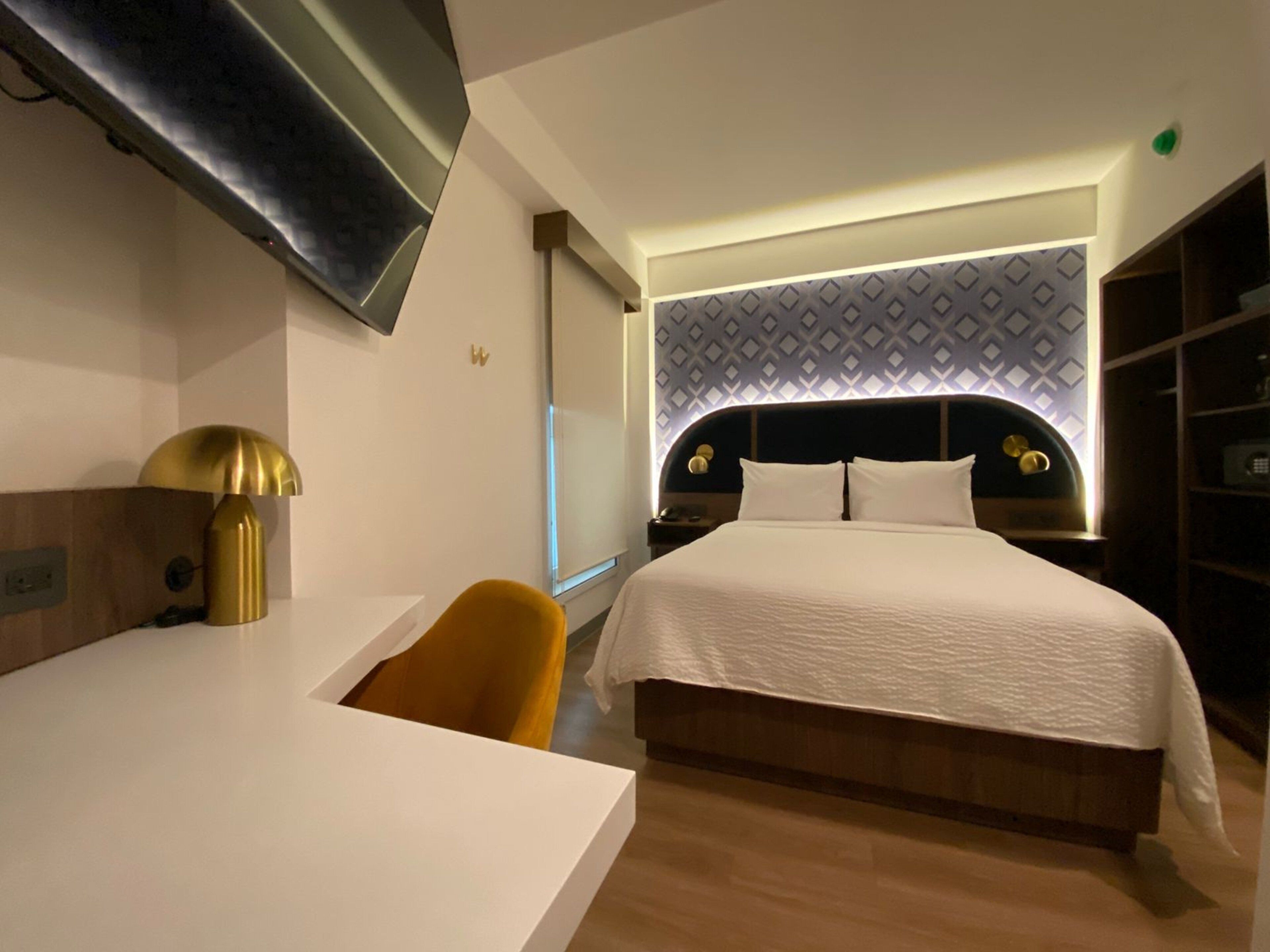 A modern hotel room with a bed, a desk, a chair, and a TV mounted on the wall.