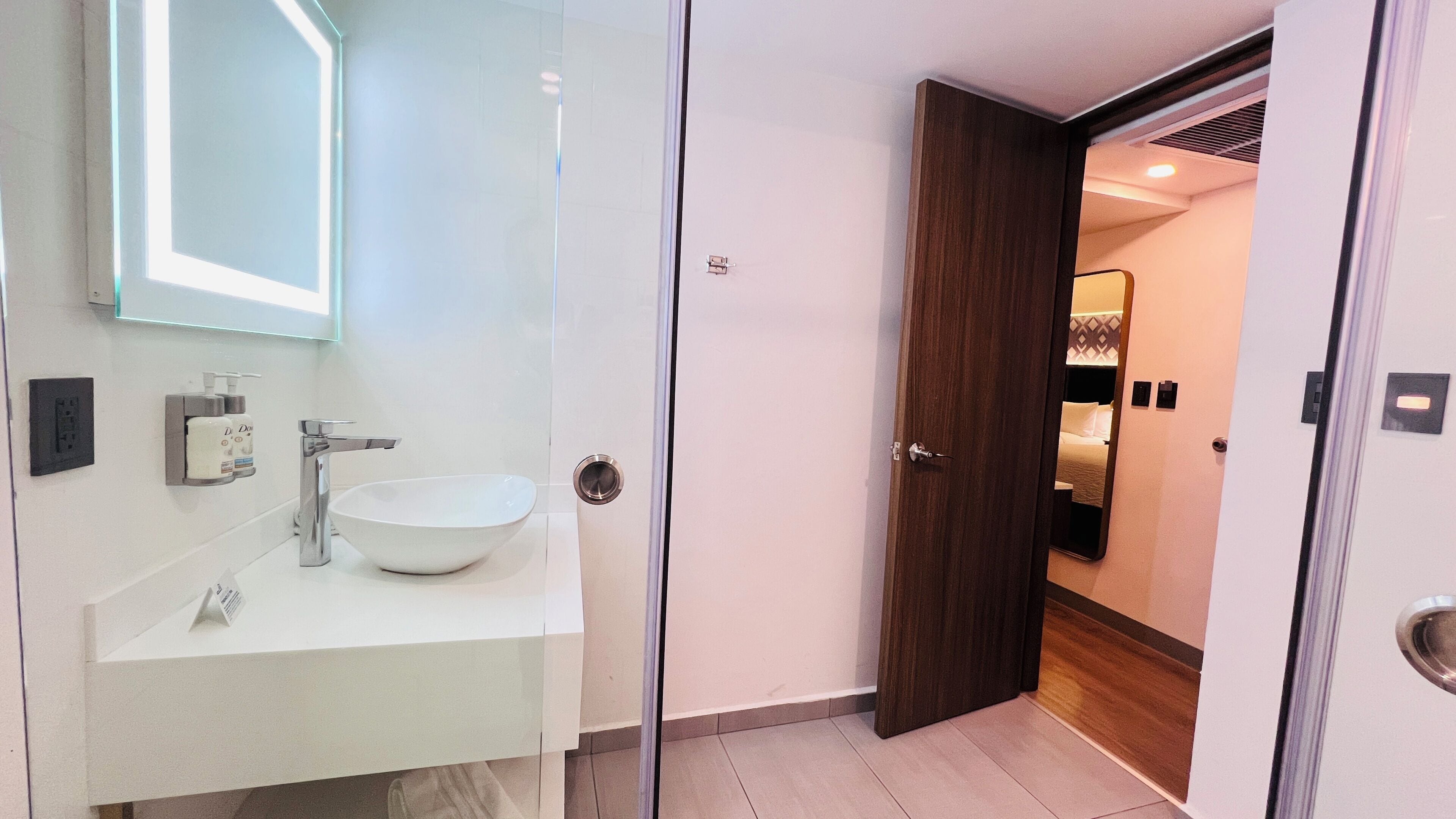 Une salle de bain moderne avec un grand lavabo blanc, un miroir et une porte donnant sur une chambre.