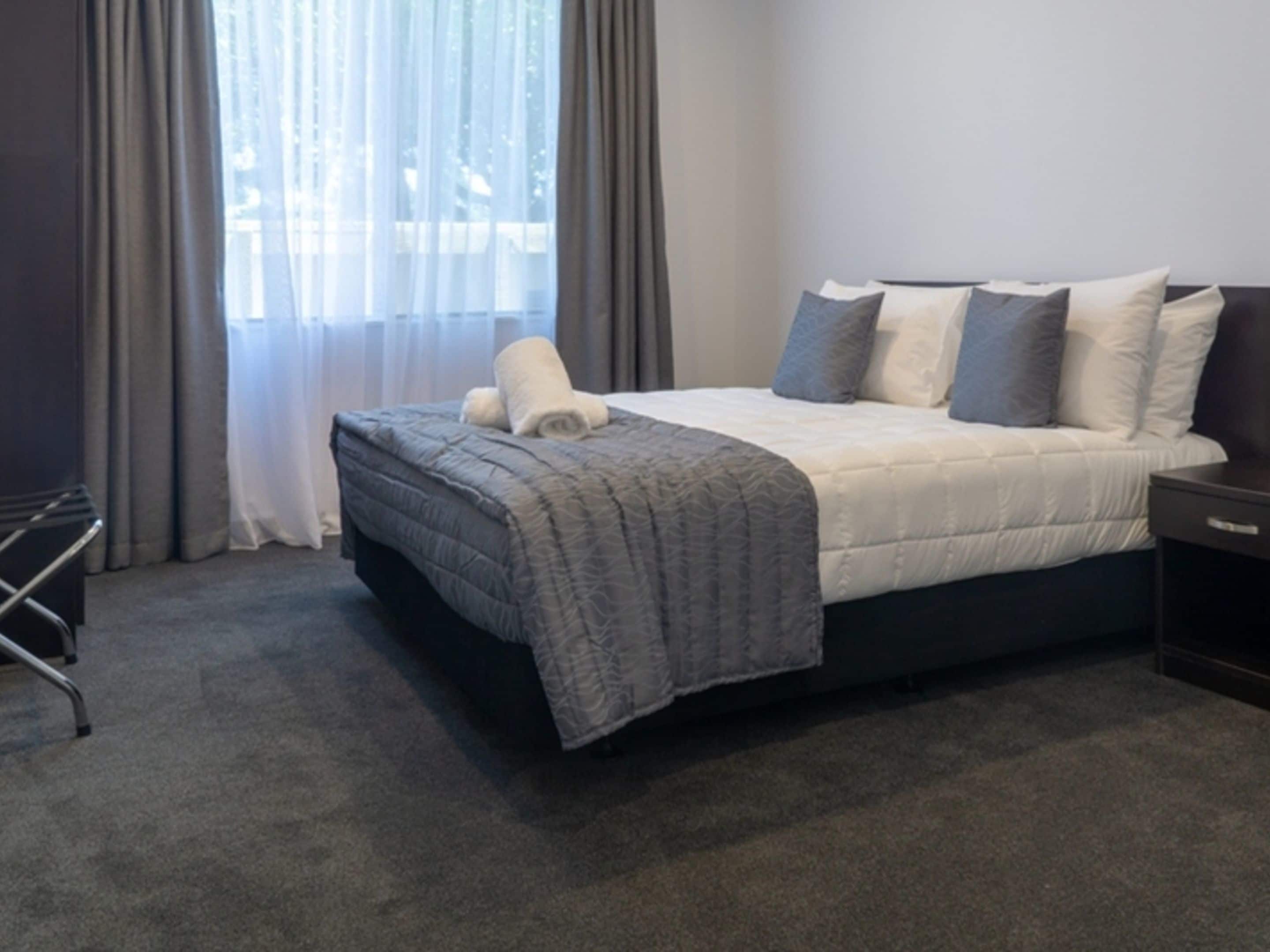 Ein ordentlich gemachtes Bett mit einer grau-weißen Decke, einem aufgerollten weißen Handtuch und mehreren Kissen in Grau- und Weißtönen. Rechts neben dem Bett befindet sich ein dunkler Nachttisch.
