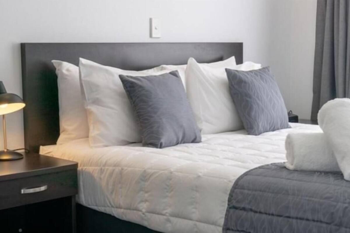 Una cama bien hecha con almohadas, una lámpara de noche y una toalla enrollada sobre una mesita de noche oscura.