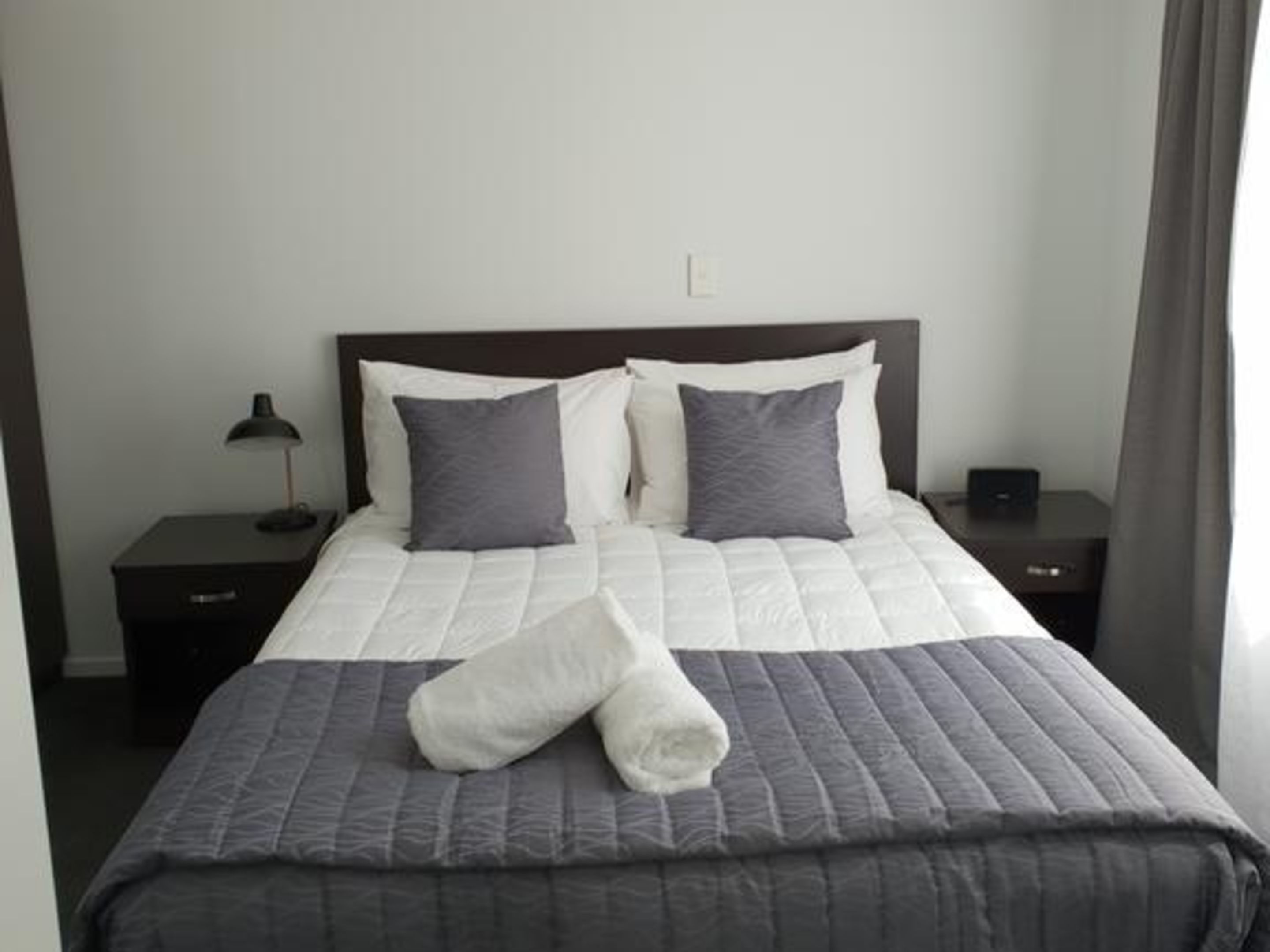 Una cama bien hecha con colcha gris y almohadas, una lámpara de noche y un pequeño dispositivo electrónico en la mesita.