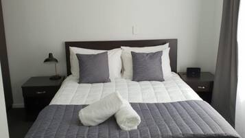 Una cama bien hecha con colcha gris y almohadas, una lámpara de noche y un pequeño dispositivo electrónico en la mesita.