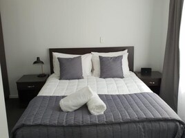 Una cama bien hecha con colcha gris y almohadas, una lámpara de noche y un pequeño dispositivo electrónico en la mesita.