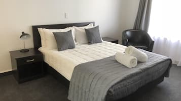 A neatly made bed with a grey blanket and pillows, a bedside lamp, a chair, and a window with curtains.