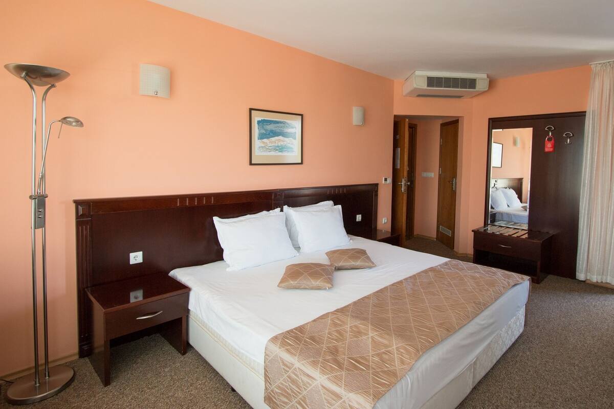A hotel room with a large bed, a nightstand, a floor lamp, a painting on the wall, and an air conditioning unit.