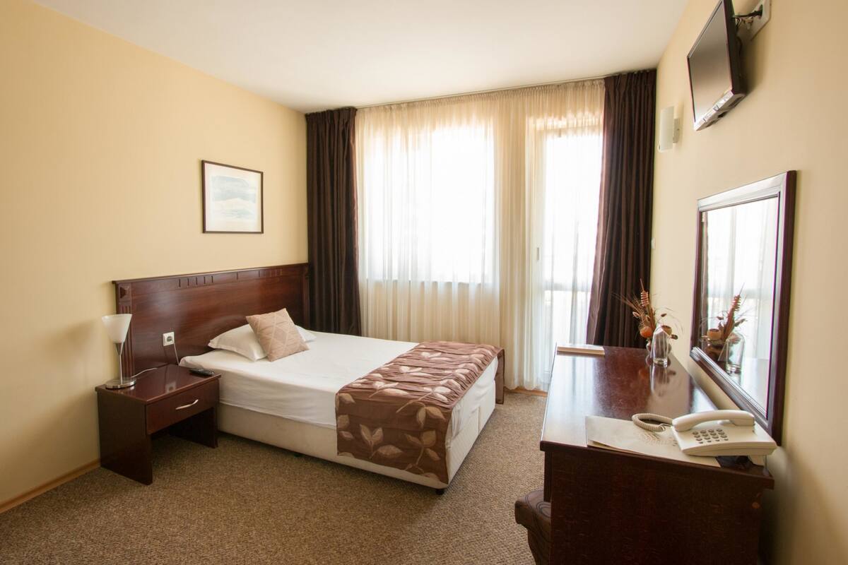 A hotel room with a bed, bedside table, lamp, desk with a telephone, and a mirror.