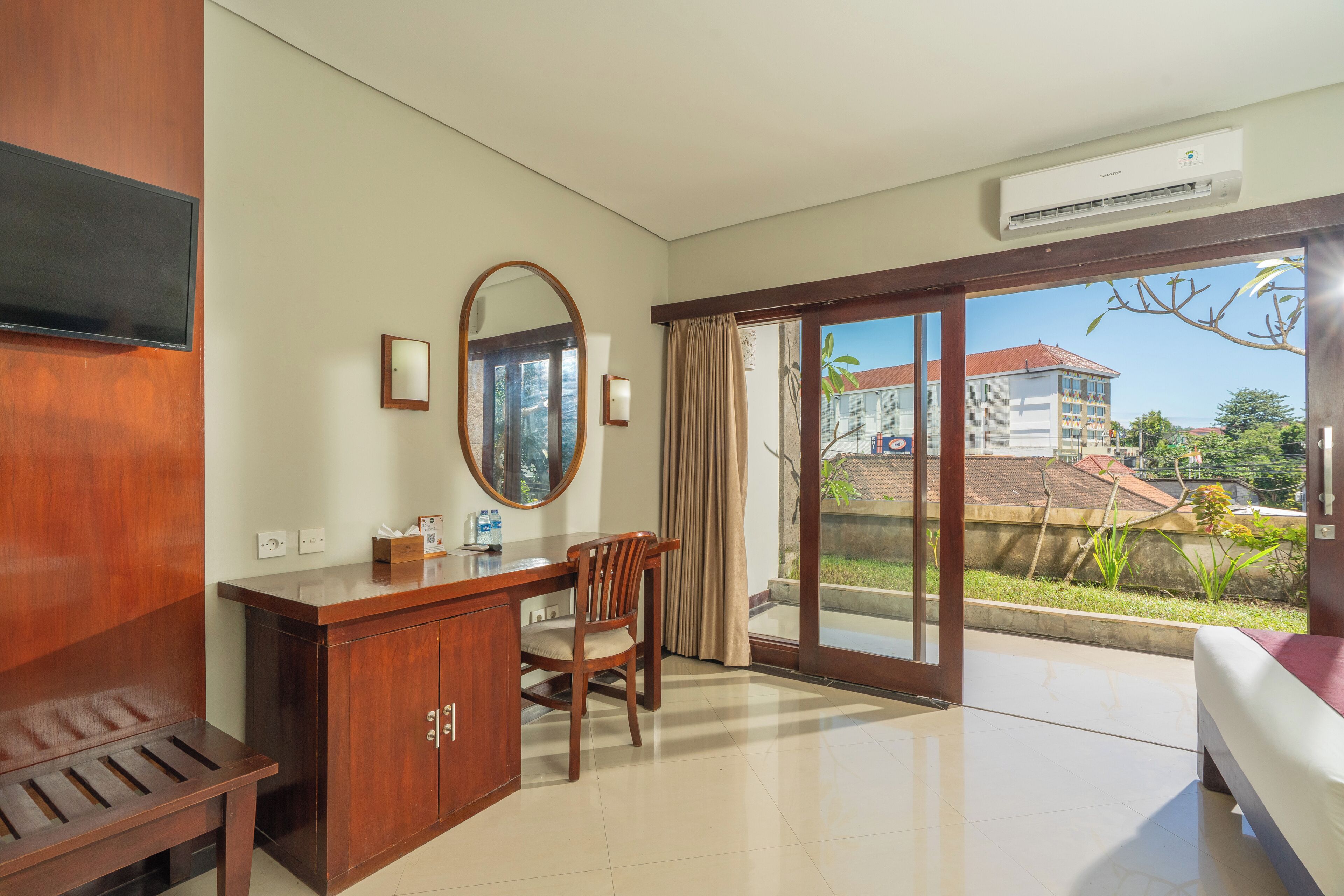 A hotel room with a wooden desk, a chair, a bed, a large mirror, and a sliding glass door leading to a balcony with a view of buildings.