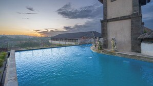 A swimming pool with a fountain, a stone building with a clock, and a view of a distant landscape during sunset. - Nirmala Heritage Hotel (Jimbaran)