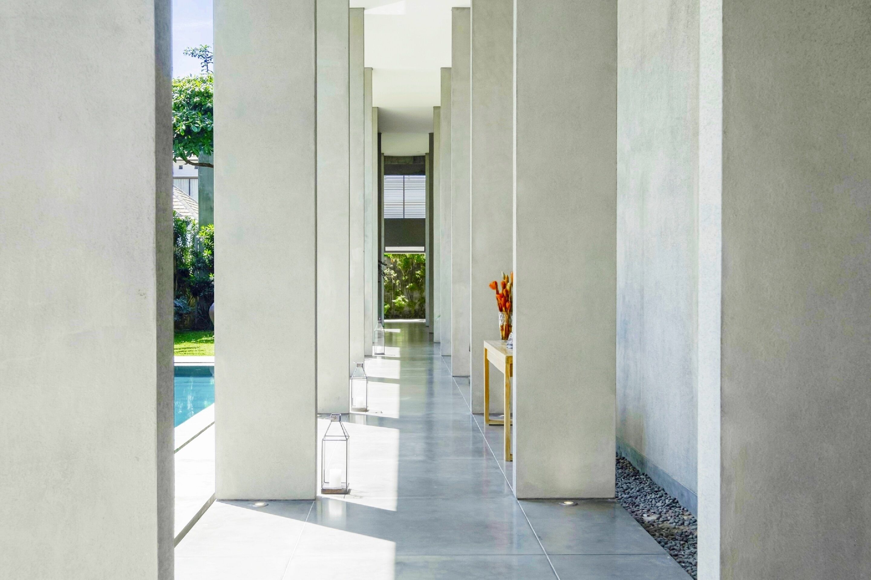 Un couloir moderne donnant sur la piscine, avec des murs en béton et une petite table surmontée d’un vase.