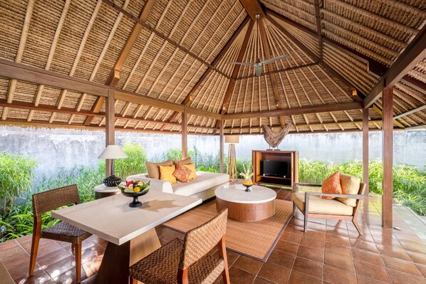A covered outdoor seating area with a thatched roof, a sofa, a TV, and a dining table with chairs.