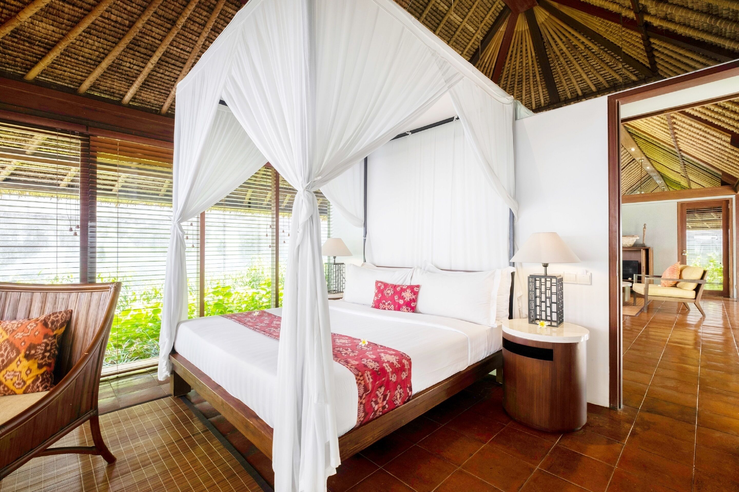 Una cama con dosel y cortinas, ropa de cama blanca, una mesita de noche con lámpara y vistas al exterior con vegetación.