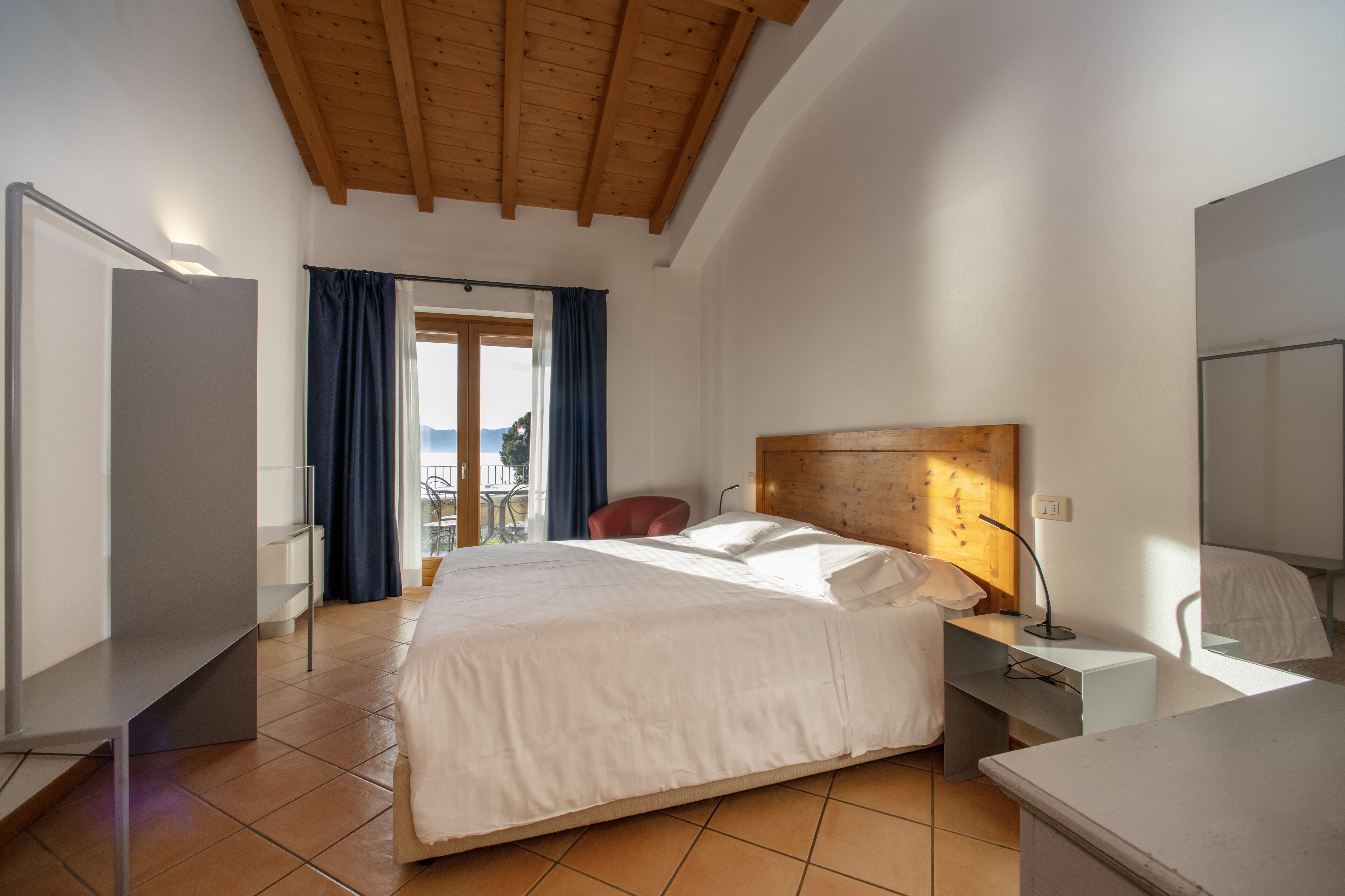 A bedroom with a large bed, a wooden ceiling, a nightstand, a desk, and a window with curtains.