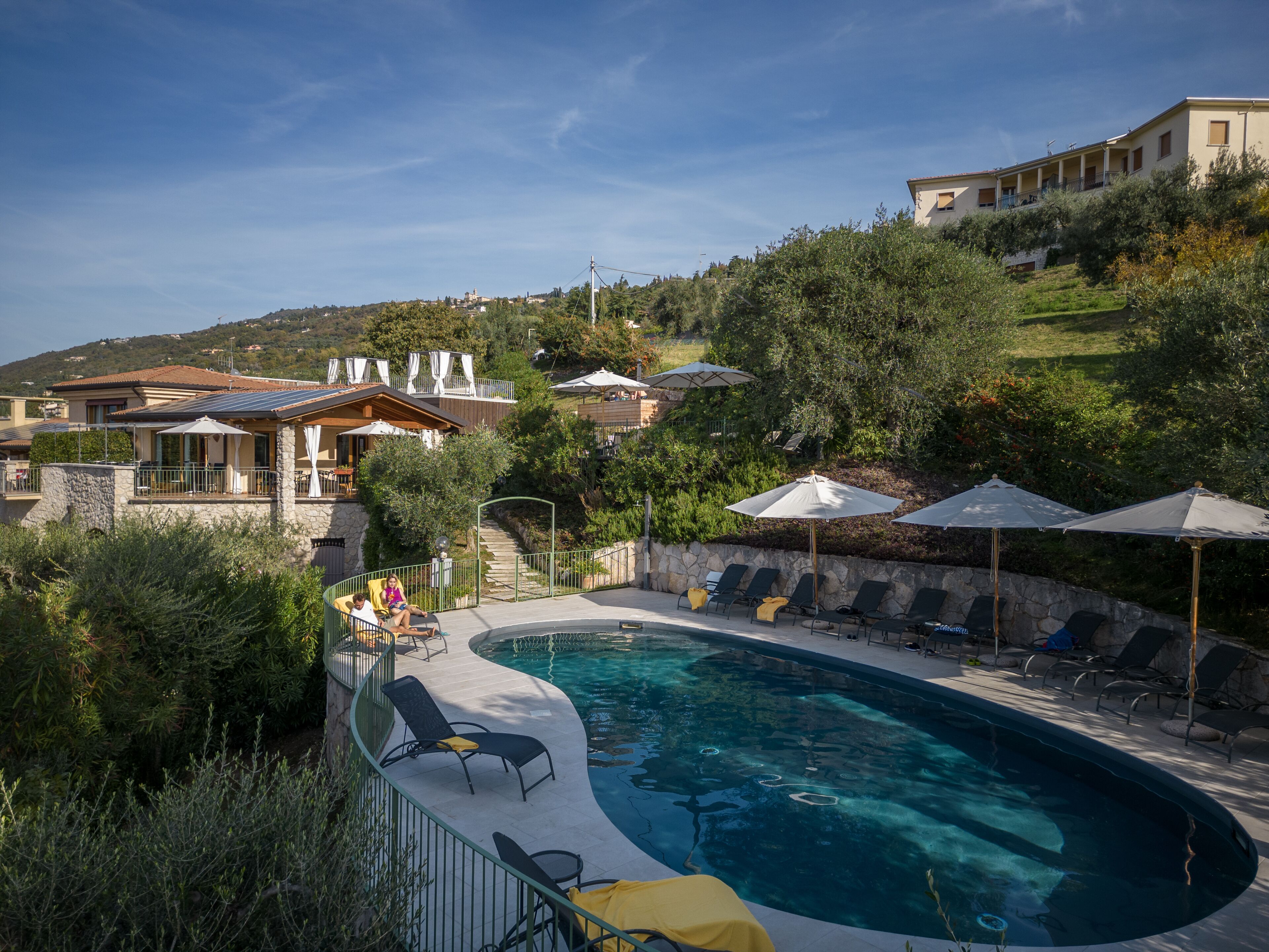 Piscina con sillones de descanso y sombrillas, rodeada de vegetación y edificios.