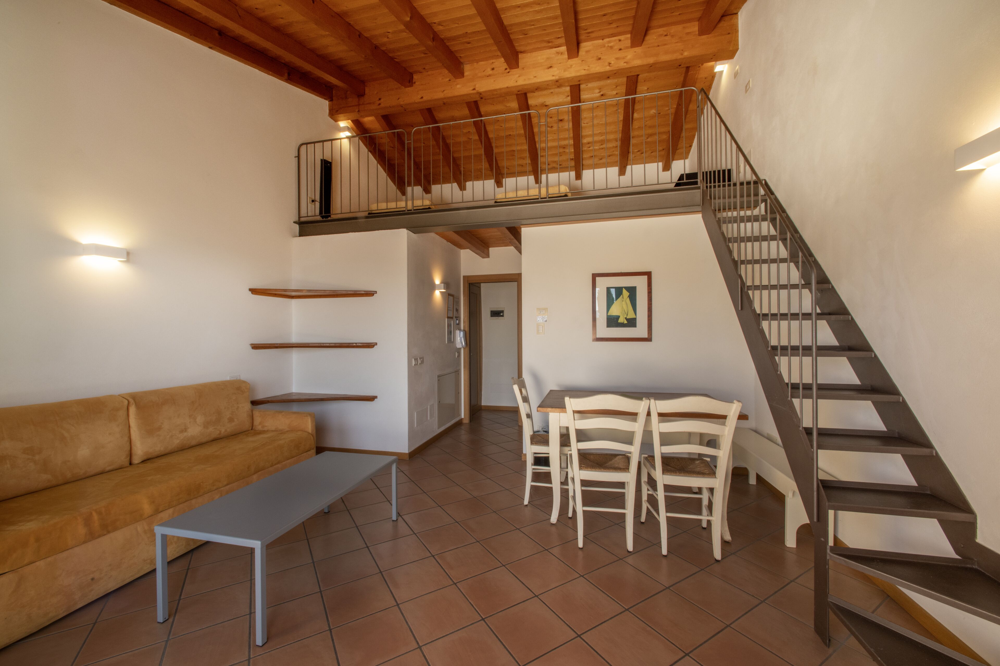 A spacious living area with a high ceiling, a wooden staircase, a sofa, a dining table with chairs, and a small balcony.