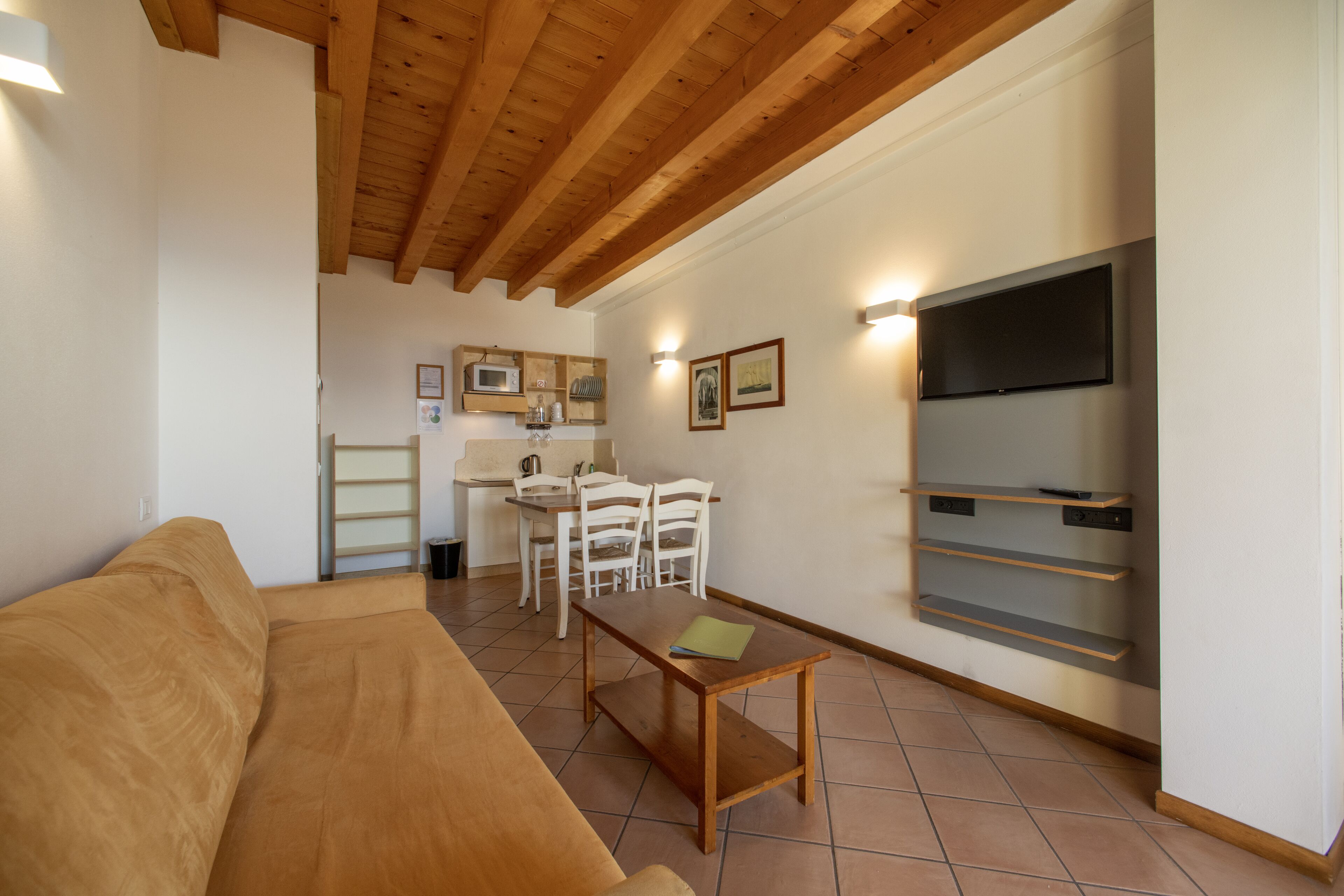 Sala de estar con techo de madera, un sofá, una mesa de centro, un televisor y una zona de cocina con microondas y sillas.