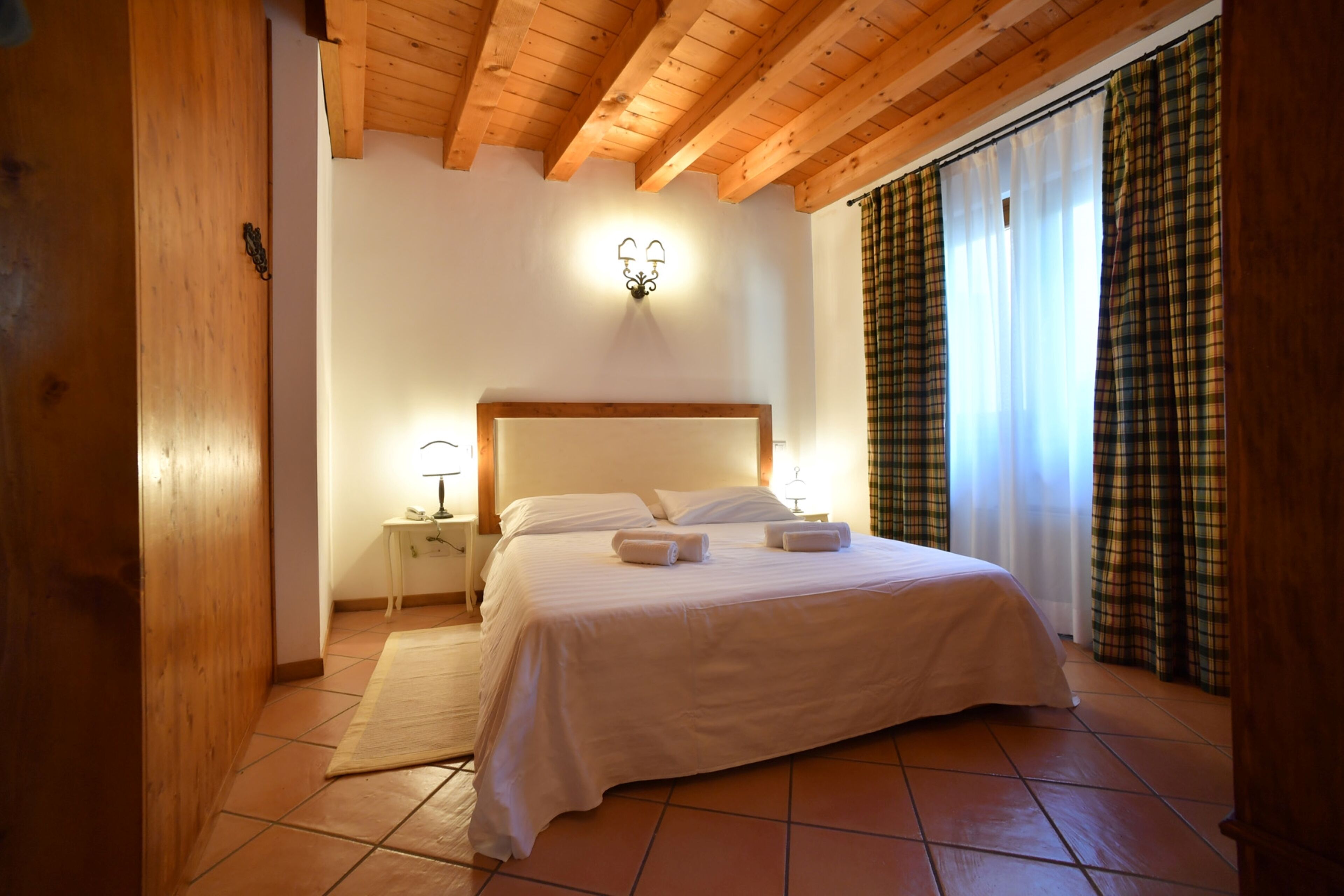 A bedroom with a wooden ceiling, a bed with white bedding, a nightstand with a lamp, and a window with curtains.