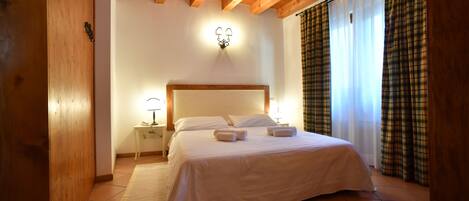 A bedroom with a wooden ceiling, a bed with white bedding, a nightstand with a lamp, and a window with curtains.