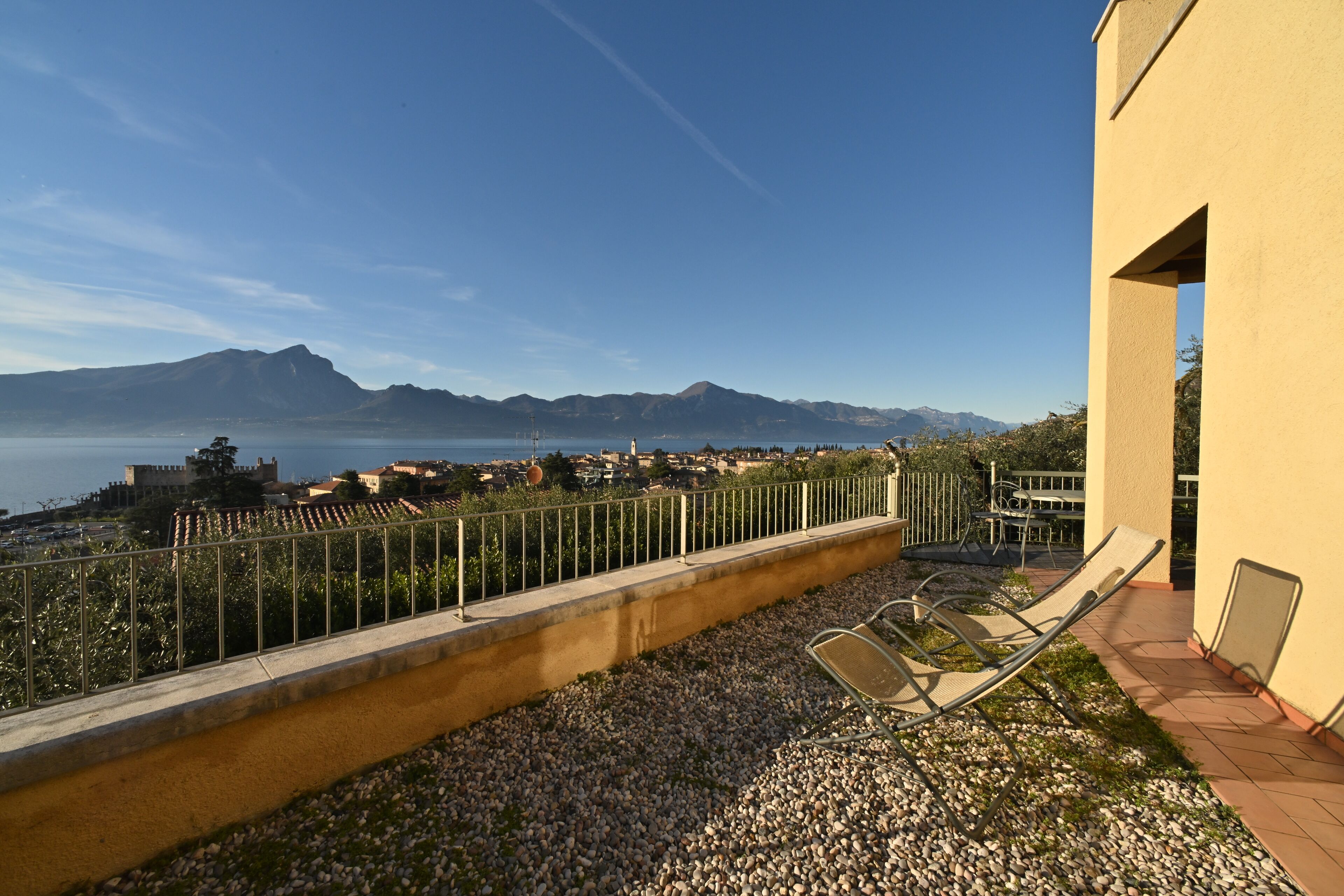 A balcony with a view of the sea and mountains, a railing, and two lounge chairs.