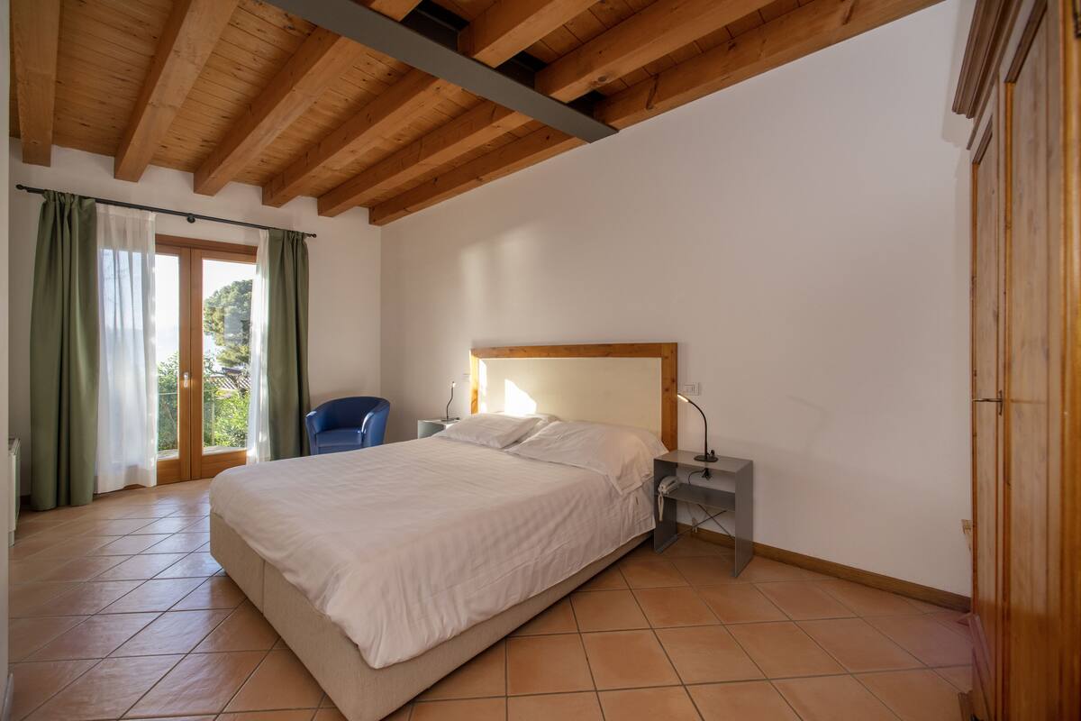 A bedroom with a large bed, wooden ceiling beams, a nightstand, a chair, and a view of greenery through a window.