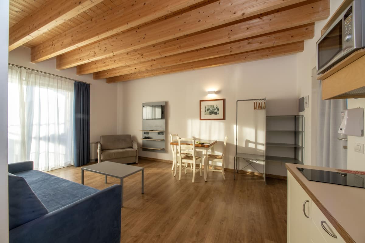 A modern living room with a wooden ceiling, a sofa, a dining table with chairs, a kitchen area with a microwave and refrigerator, and a large window with curtains.