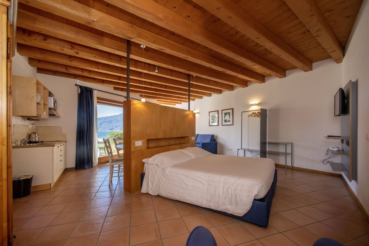 A bedroom with a large bed, wooden ceiling beams, a view of the outdoors, a small kitchenette, and a bathroom with a mirror and towel rack.