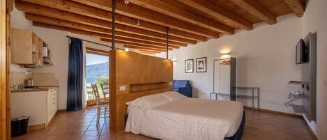 A bedroom with a large bed, wooden ceiling beams, a view of the outdoors, a small kitchenette, and a bathroom with a mirror and towel rack.