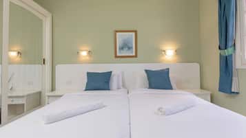 Quarto duplo com duas camas, roupa de cama branca e almofadas azuis. Há um quadro na parede e duas luzes fixadas na parede.