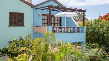 A light blue building with a balcony, surrounded by lush greenery and a clear sky.