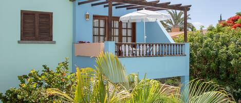 A light blue building with a balcony, surrounded by lush greenery and a clear sky.