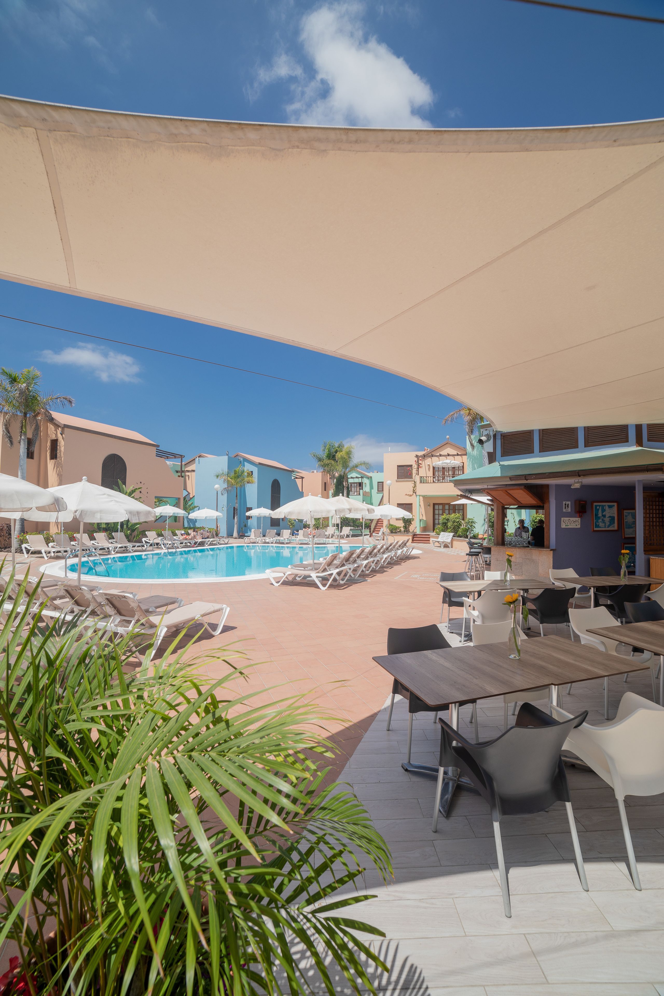 Ett område vid poolen med solstolar och parasoller, en pool och en byggnad med en blå fasad.