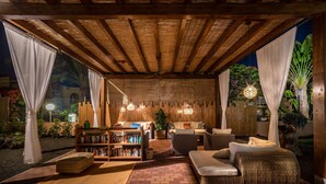 A cozy outdoor lounge area with a wooden ceiling, comfortable seating, a bookshelf, and a well-lit ambiance.
