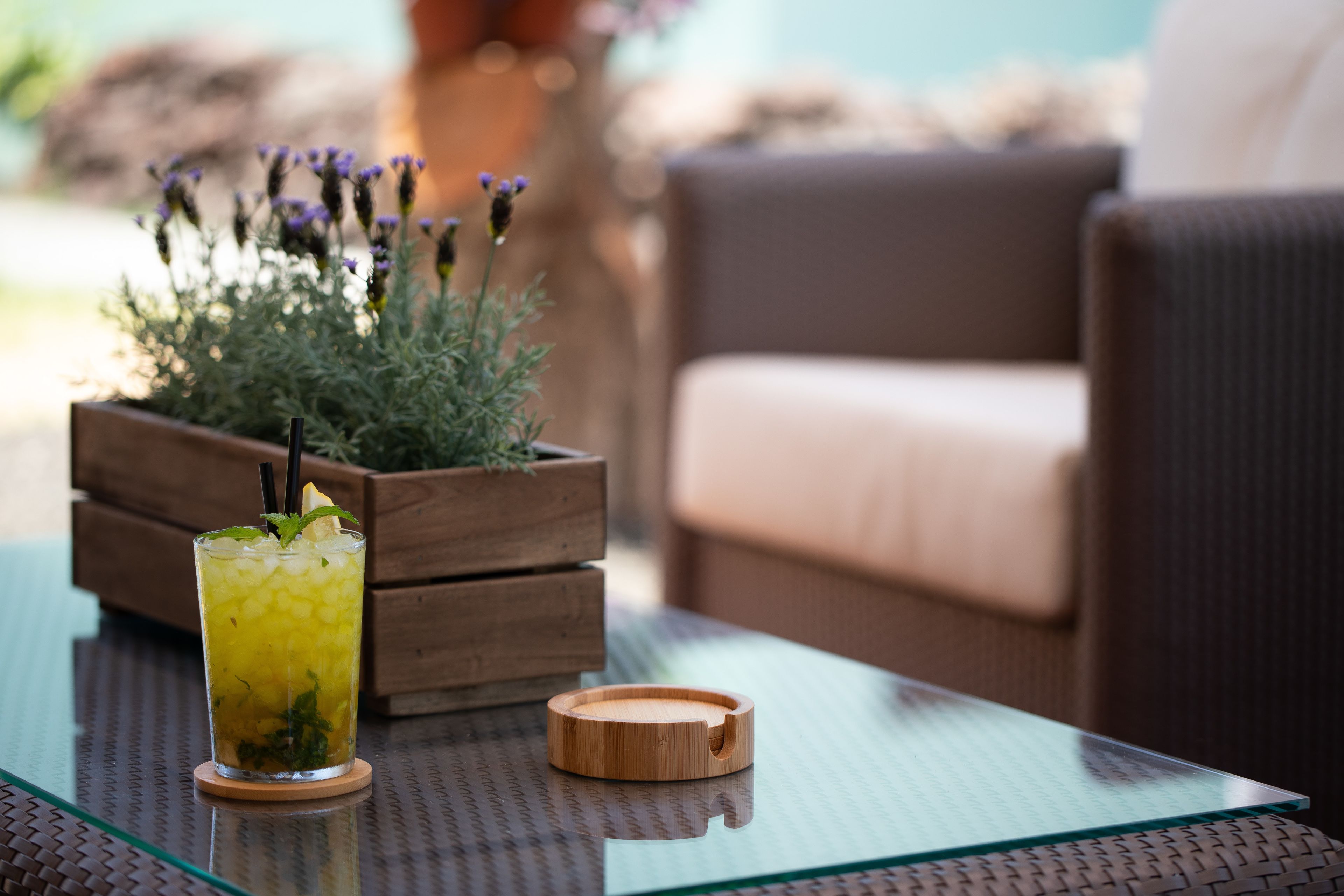 Un vaso de bebida helada sobre una mesa de vidrio con un posavasos de madera, una maceta de madera con flores y un sofá marrón al fondo.