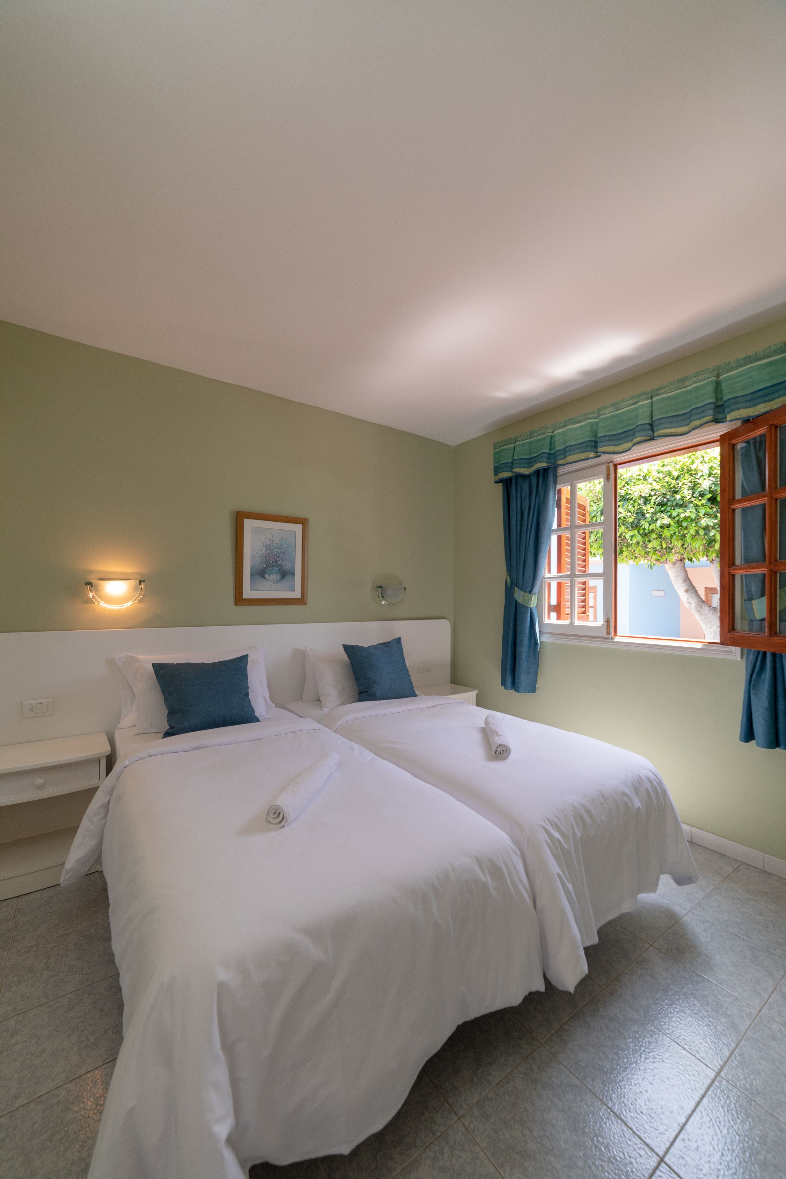 A room with two beds, a nightstand, a wall-mounted light, a window with curtains, and a framed picture on the wall.