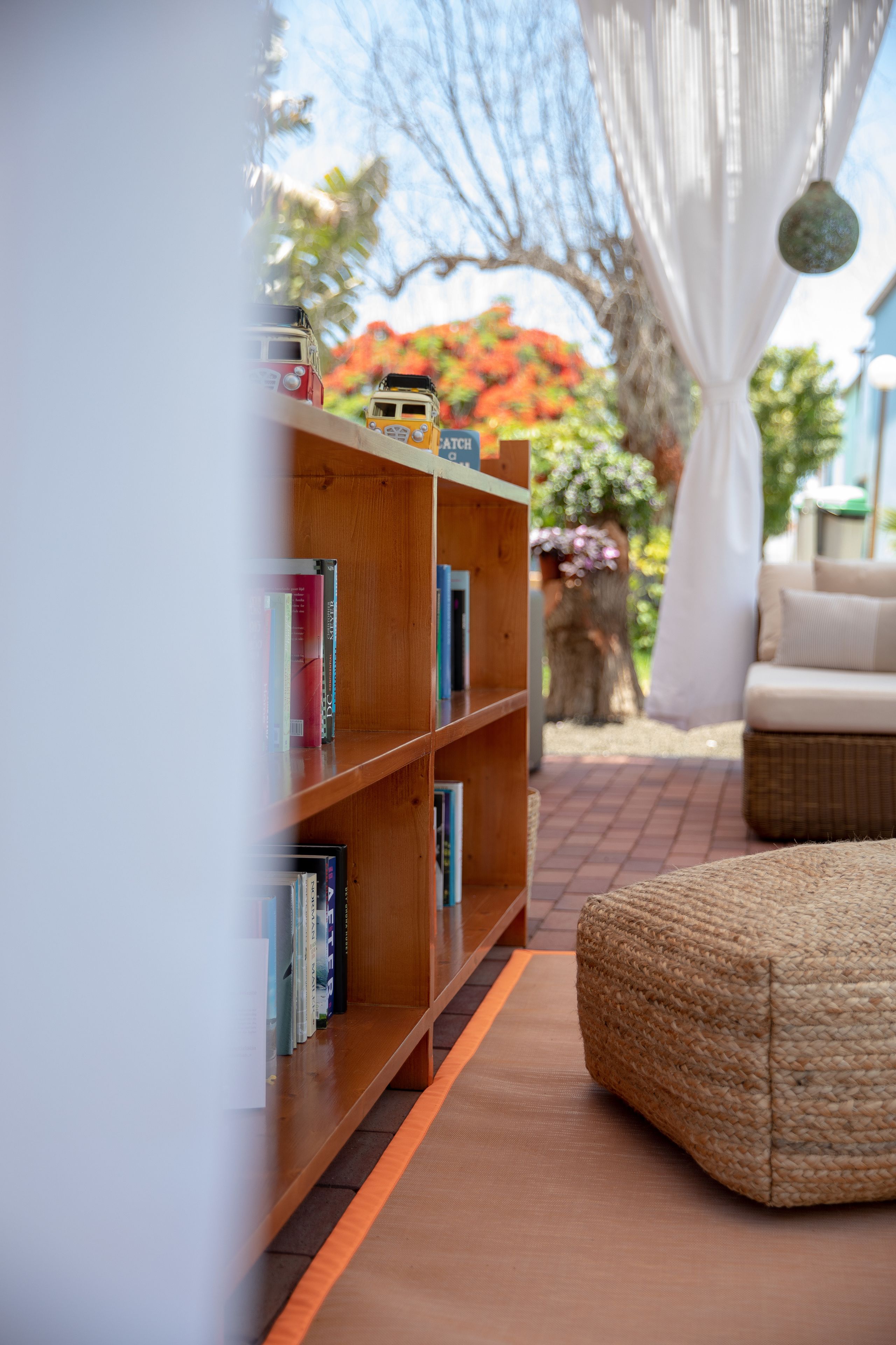 A wooden bookshelf with books, a wicker ottoman, and a view of a garden with a tree and flowers.