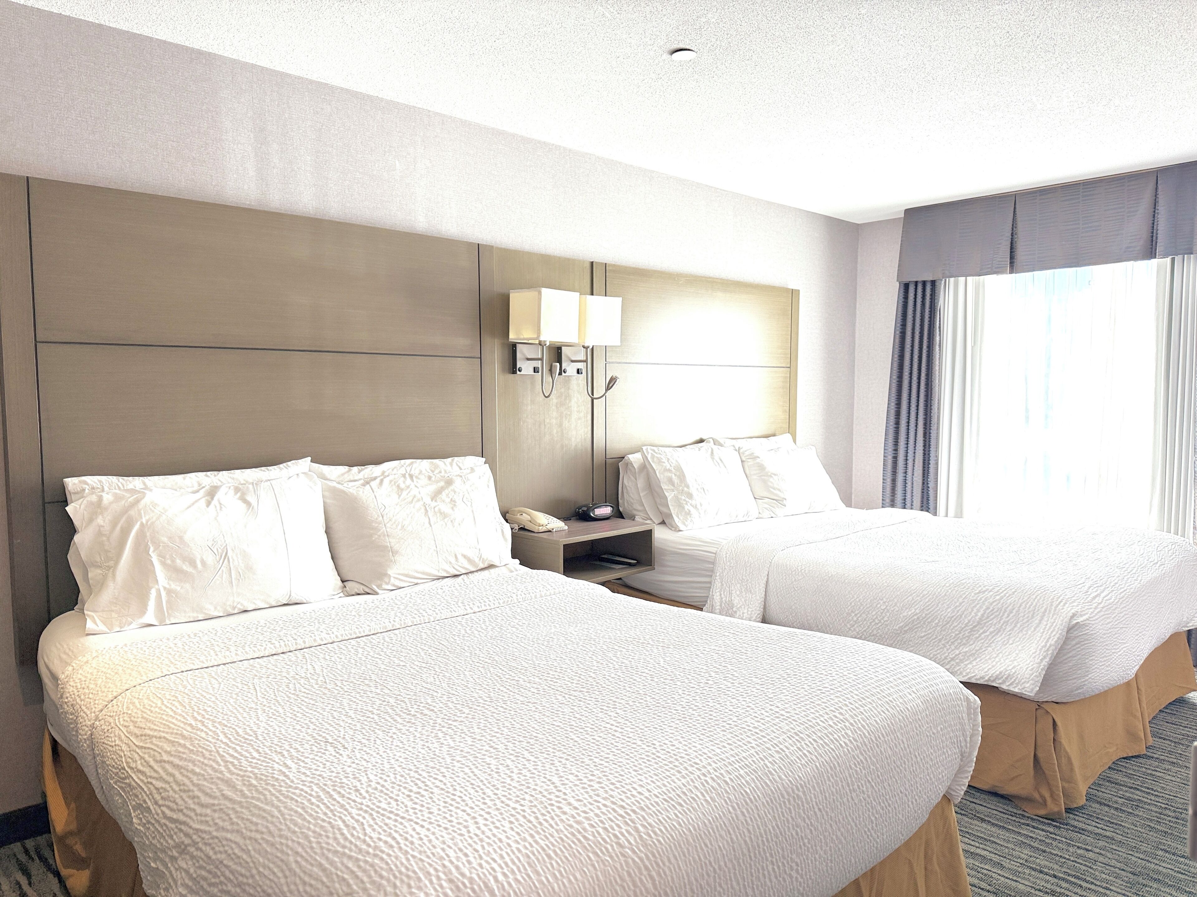 A hotel room with two beds, a nightstand with a lamp, and a window with curtains.