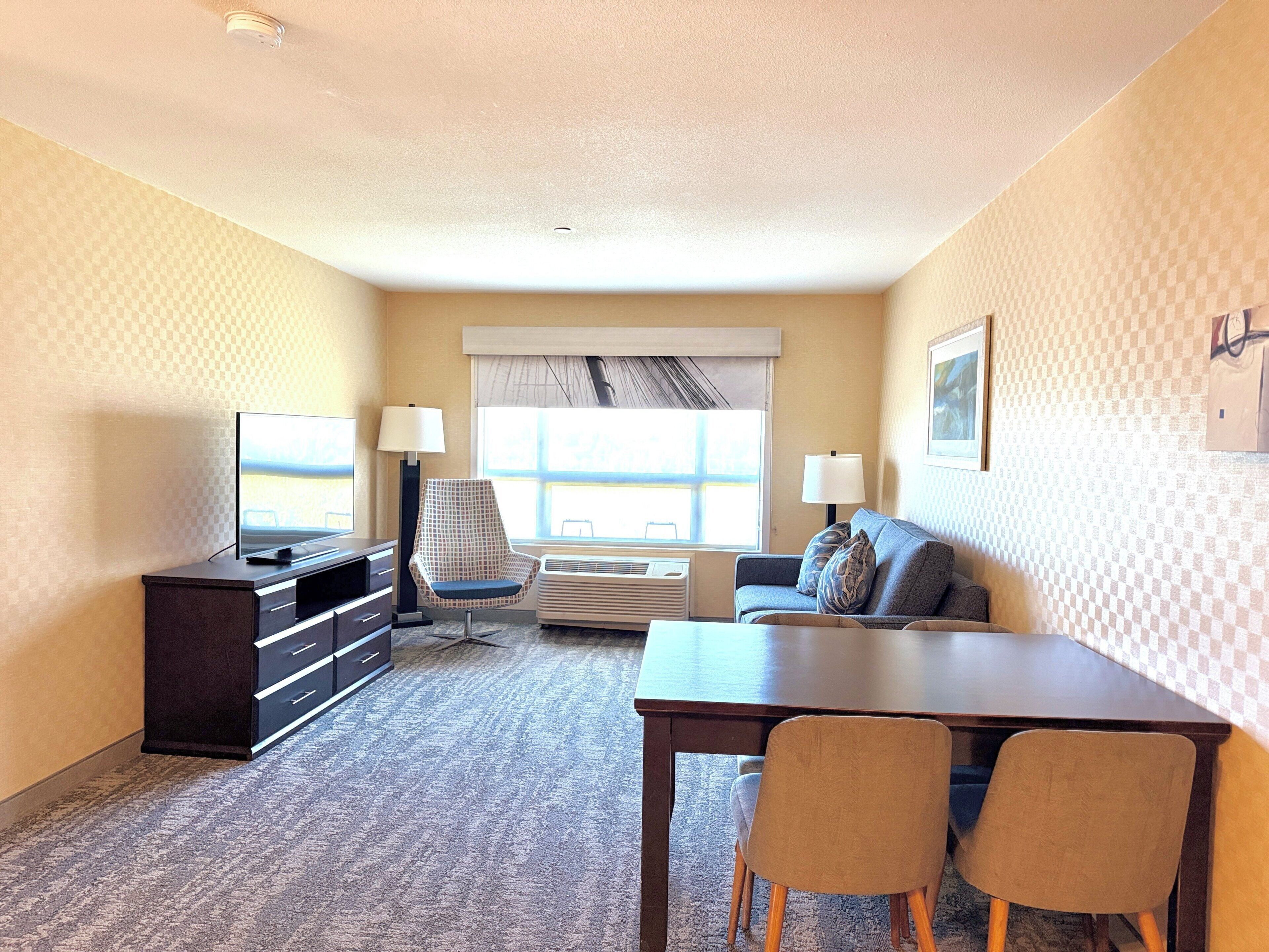 A hotel room with a TV, two chairs, a table, a sofa, and a window with curtains.