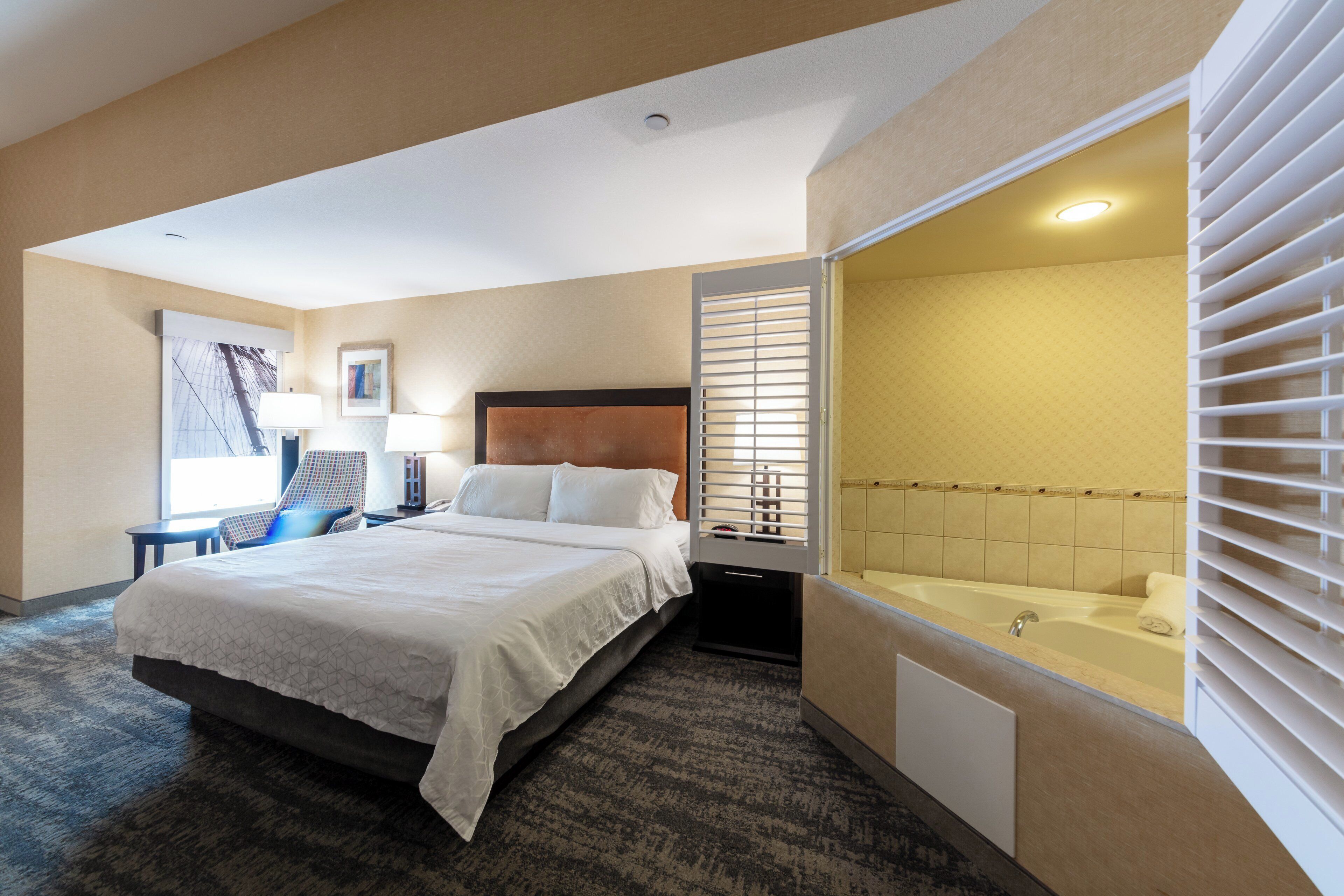 A hotel room with a bed, a desk, a chair, a window, and a bathroom with a bathtub.