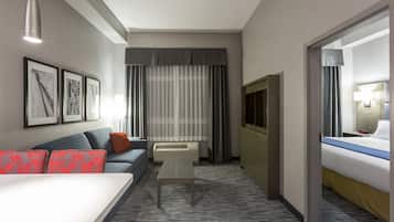 A hotel room with a sofa, a small table, a bed, a TV, and a window with curtains.