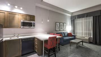 A compact hotel room with a kitchenette, a sofa, a dining table, and a large window with curtains.