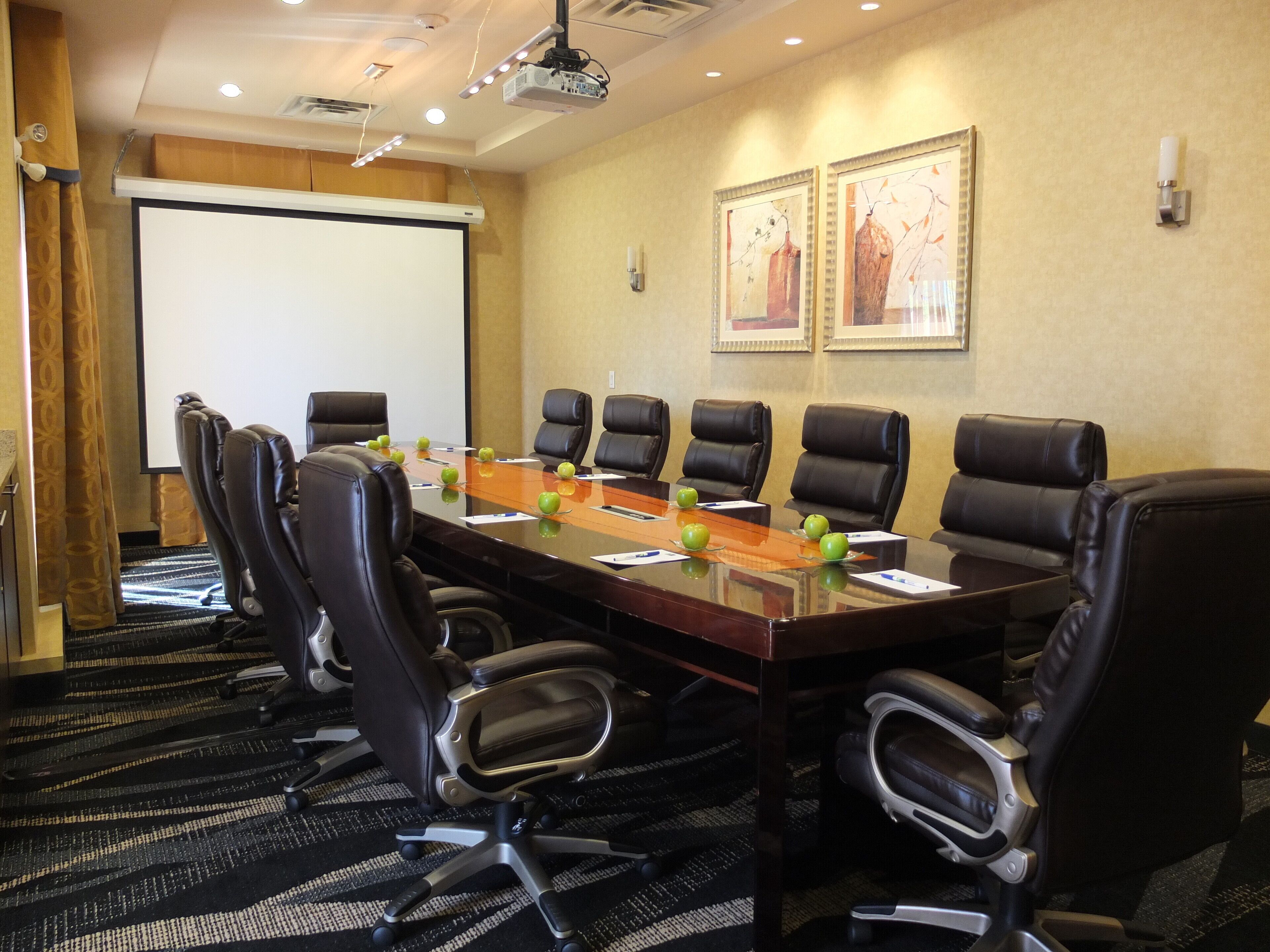 A conference room with a long table, black leather chairs, a projector screen, and two framed artworks on the wall.