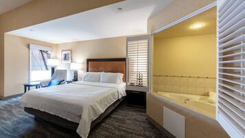 A hotel room with a large bed, a sitting area with a chair and table, a bathroom with a bathtub, and a large window with blinds.