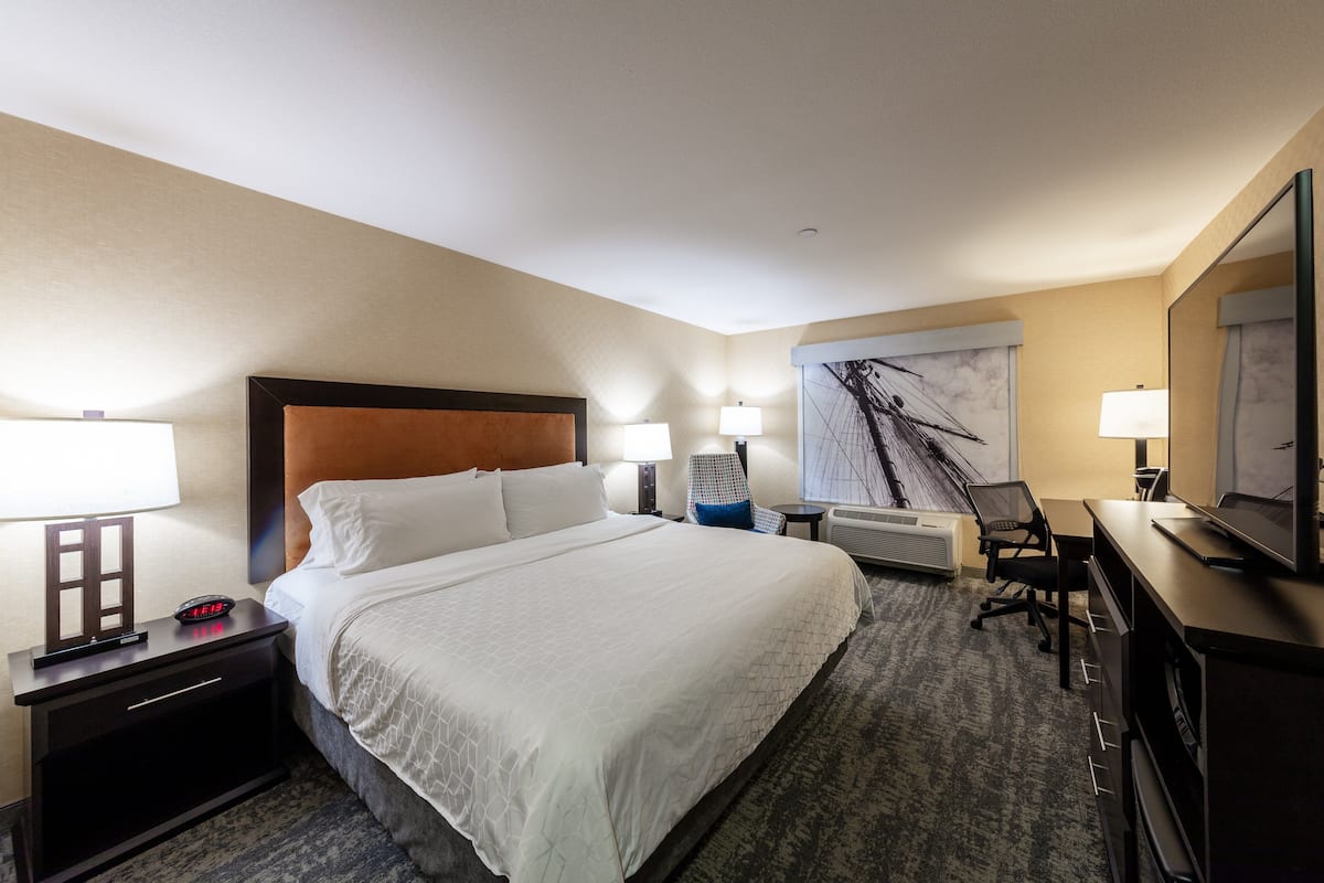 Une chambre d’hôtel avec un grand lit, des tables de chevet avec des lampes, un bureau avec un ordinateur et une grande fenêtre donnant sur un pont.