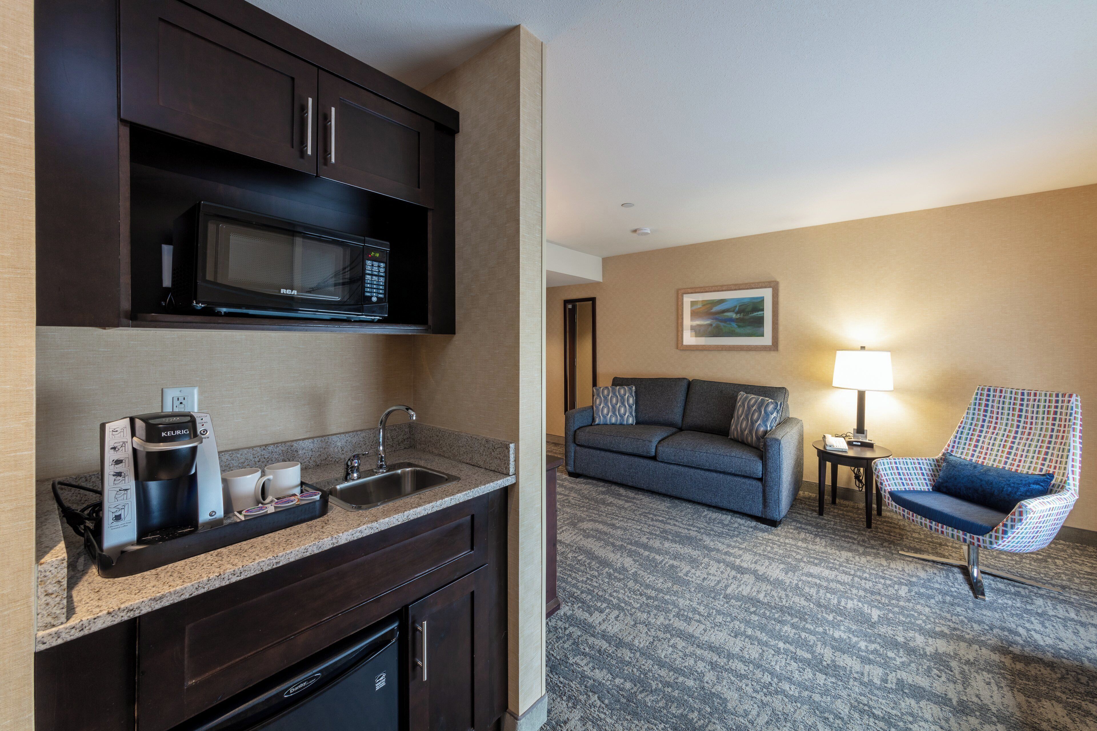 A compact hotel room with a kitchenette, a microwave, a coffee maker, a sofa, a patterned rug, and a small table with a lamp.