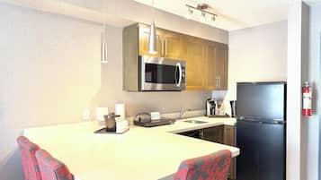A compact kitchen with a microwave, refrigerator, and a coffee maker. There are two chairs with a floral pattern, a small island with a kettle, and a fire extinguisher mounted on the wall.