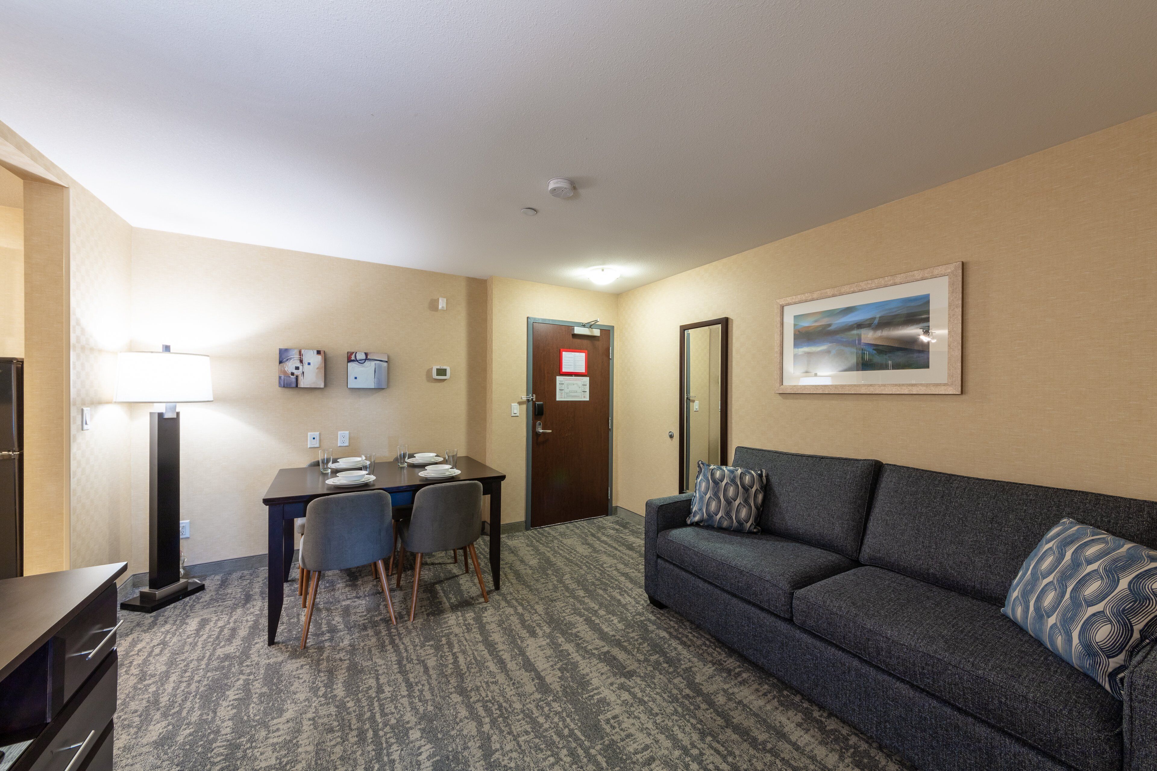 Une chambre d’hôtel avec un canapé gris foncé, une petite table avec des chaises, une lampe et un tableau encadré au mur.