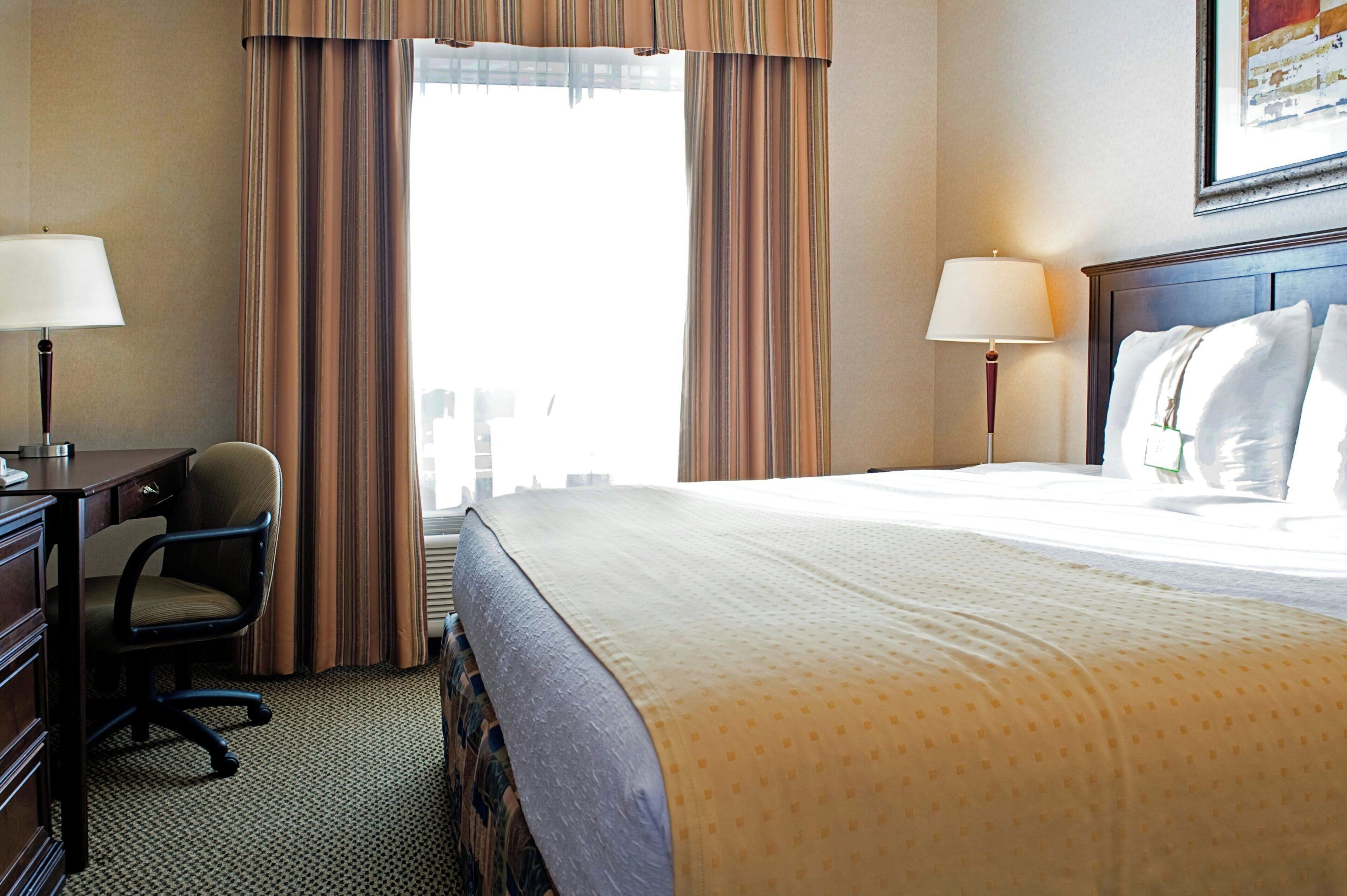 A hotel room with a bed, two bedside lamps, a desk with a chair, and a window with curtains.